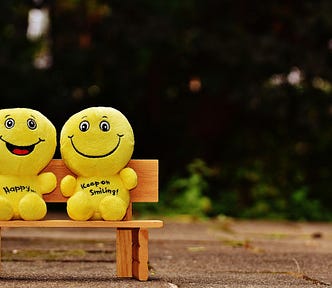 Photo of two yellow smiley faced emoji stuffies, sitting on a wooden bench outside. The one on the left has an open-mouthed smile and says “HAPPY…” on the front, and the other says, “KEEP ON SMILING!”