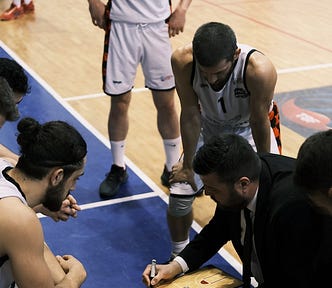 Basketball players on the sideline watching as a coach outlines a plan on a clipboard.
