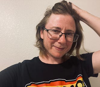 Author is wearing a t-shirt and black framed glasses. He is holding his hair back and has the beginnings of a beard. He’s smiling just a little.