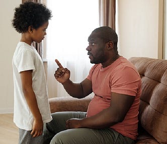 A father sitting on the sofa talking to his son. His finger pointing at his son who is standing in front of him.