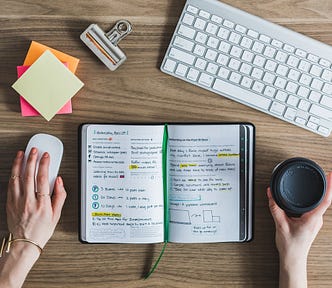 A desktop with a journal, keyboard, mouse, coffee cup, and a pair of hands