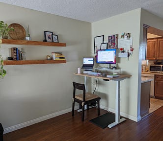 A corner of a room. On one corner are two wooden shelves with books and a plant on them. On the other corner is a sit/stand desk with photos on the wall, a large monitor, a laptop on a stand, and a keyboard, agenda, mouse, and water bottle.