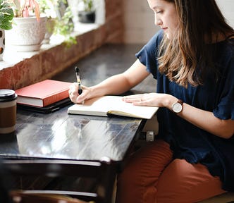 A woman writes in her journal at a table with plants, coffee and books.