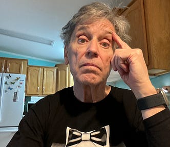 A man with gray hair and light skin is sitting in a kitchen, wearing a black T-shirt with a printed bowtie design. He has a slightly concerned or puzzled expression, resting his forehead on one hand. Behind him, there are wooden kitchen cabinets, a white refrigerator with colorful magnets, and a microwave. The lighting is natural, coming from an unseen source.