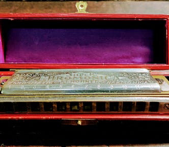 An antique metal Chromonica harmonica sitting in a wooden box with a purple internal lining and red trim.