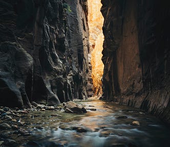 A river pebbled with stones runs through a large canyon. The light bleeds through.