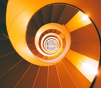 A spiral staircase viewed from below, along the middle of the spiral, with the underside of the steps resembling an upside-down staircase, warmly lit by pairs of lamps on the underside of steps on the left and right sides and leading up into a white grate.