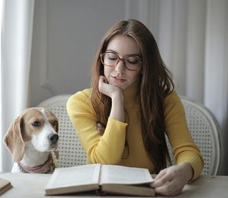 A woman with long brown hair and glasses wearing a yellow turtleneck sits at a table reading an open book. A dog sits next to her and looks on.