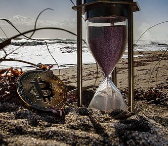 An hourglass and a bitcoin coin are on the sand on a beach. Image accompanying the Harlow Journey story, “Can the Government Shut Down Cryptocurrency?”