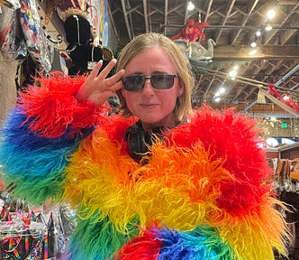 Author is wearing a rainbow feathered costume in a costume store. An elephant is peeking out from behind his head in the background. Other colorful costumes and masks can be seen on the wall behind him. There are lights along the wooden ceiling.