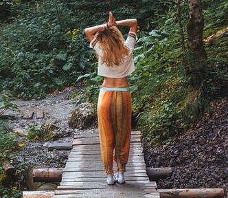 Woman standing on a wooden bridge with arms raised above her head, hands joined. The background is nature, greenery and trees.