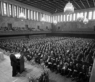 A large ballroom filled with people seated in rows, listening to a man at lectern.