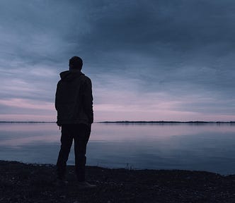 Man stood by a lake of water at dusk, staring into the distance.