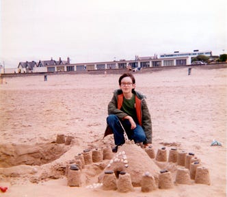 Picture from around 1980, of me around 12 years old on a quiet beach, wearing a coat, crouching down near a small mound of sand surrounded by several sand pies. I was wearing a winter coat, so it was probably cold and windy.