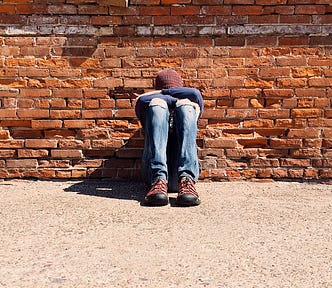 A person in blue jeans. brown boots and beanie sitting on the ground with their back against a brick wall. They have their feet on the ground and their knees up. Their arms are folded across their knees and their head is resting on their arms so their face is hidden.
