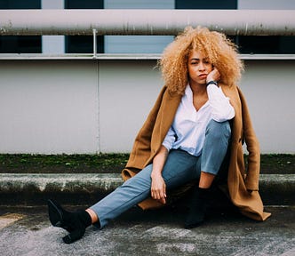 woman wearing tan coat over her shoulders sitting on ground in parking lot