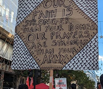 Photo of a placard from a protest with text surrounded by Palestinian keffiyeh material reading ‘Our faith is stronger than your bombs. Our prayers are stronger than your army’.