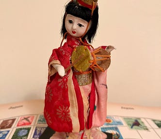 A Japanese doll in traditional costume holding a small drum and standing on a stamp album opened at pages headed ‘Japan’.