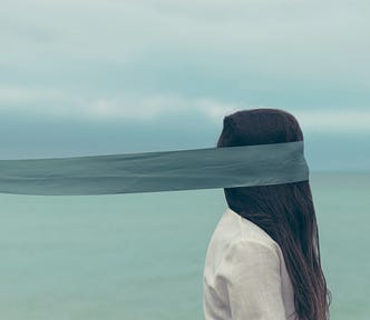 person facing the right with a wide teal ribbon covering their face and trailing far behind them out of the image, person’s head is covered in long, straight dark hair falling onto a white shirt, opaque sea or lake and cloudy sky in the background