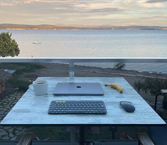 Agean Sea view from a balcony of Burla Han Hotel in Gülbahçe, İzmir, Türkiye. There is a MacBook, a keyboard, a mouse, a cup of coffee and a banana on the table.