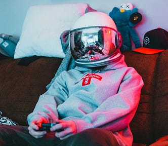 Man wearing a hoodie and an astronaut helmet plays video games in is living room after having swindled another victim on Tinder.