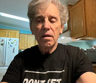 An older man with gray hair wearing a black t-shirt that has partially visible text reading “DON’T GET CAUGHT.” He is seated in a well-lit kitchen with wooden cabinets and a refrigerator in the background. The refrigerator has decorative magnets, including butterfly designs. The man appears to be looking downward with a neutral or contemplative expression.