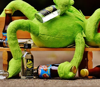 Kermit The Frog lying on wooden bench surrounded by glass bottles. Kermit appears drunk with a bottle of clear bottle presumed to be vodka in one hand and a brown bottle presumed to be beer in the other. He’s lying on his back with his mouth open and one leg hanging over the back of the bench.