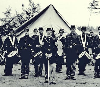 Black and white photograph of 14 men in Union uniforms standing outside a tent. Most carry drums, some carry fifes. The man standing front and center holds a sword in front of him with the point on the ground. Two of the drummers appear to be black; the remainder of the men appear to be white.