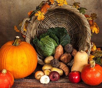 Wicker basket wreathed with orange and yellow leaves (background); fall harvest vegetables — pumpkin, squash, onions, potatoes, beets, yams, cabbage and apples (foreground)