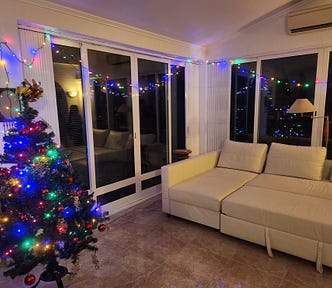 A three-foot tall Christmas tree with multicolored lights and red and gold ornaments. There are lights strung in front of large glass windows and a white couch to one side.