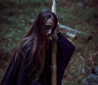 Mysterious Woman / witch with long dark hair and black robes and a shadowy face holds a staff that is partly charred, background of woodland