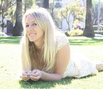 Light-skinned platinum blonde woman in her forties, laying on the grass on a sunny day in a park, facing the camera. She’s wearing a light floral dress and smiling, hands in front of her. She’s the author’s friend, the subject of the story