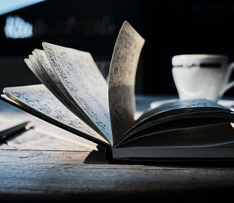 Photo of a book on a wooden table. The book is flipped open, many pages visible with handwritten text. A pen lays on the table to the left of the book, and slightly behind the book on the table. A teacup is behind the book on the right of the frame. The distant background is dark, with some light in the top left corner.