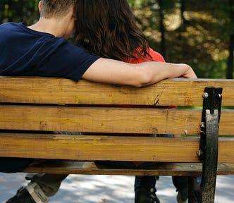 Rear view of a man and a woman sitting on a park bench. The man has his arm around the woman’s shoulders. She is leaning in, either whispering in his ear, kissing him, or simply being close.