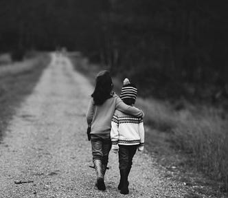 Black and white, two children walking down a road. Taller child has arm around shorter child.
