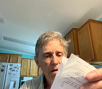 An older man with gray hair is sitting in a kitchen, looking intently at a paper receipt he is holding in his hand.