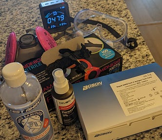 On my kitchen counter: A box of 20 RAT COVID tests, a bottle of hydrogen peroxide spray, ethyl alcohol hand sanitizer, a box of black vinyl gloves, protective goggles, elastomeric mask, carbon dioxide monitor.