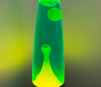 A close-up image of a green lava lamp against a gradient background transitioning from yellow to green. The lava lamp contains blobs of wax in various stages of ascent and descent, with one large drop-shaped blob suspended near the top and a round blob at the bottom, creating an interesting visual effect.