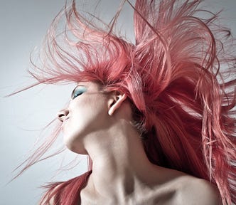 A young woman who has tossed her head to the side, flipping her pale pink hair wild in the air, against a gray background.