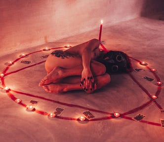 A naked woman, lying on her side, curled in a fetal position, in the centre of an occult symbol ringed with candles and tarot cards.