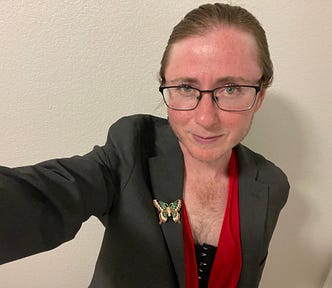 The author has his hair pulled back into a ponytail. He’s wearing black-rimmed glasses and a grey suit with a butterfly pin on it. Under the suit, you can see a bright red shirt hanging low over a black undershirt. Above the shirts, plenty of chest hair is showing. He’s smiling with a smirk that makes him look like a queer, trans masculine Mona Lisa.