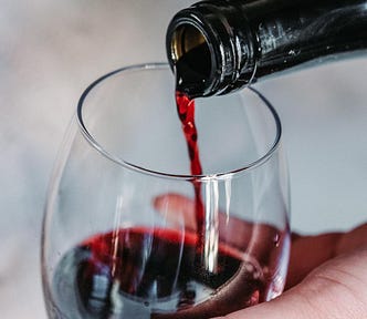 A relatively close image of red wine being poured from a wine bottle into a wine glass being held by a hand.