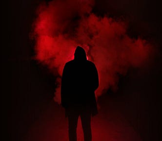 A hooded figure, standing in foggy darkness, silhouetted by a red light behind him.