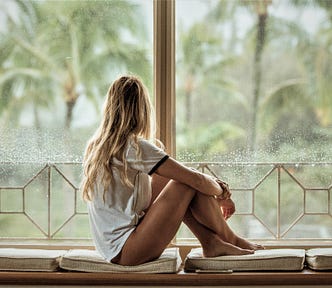 back view of girl with long blonde hair wearing white t-shirt sitting on cushion looking out a window