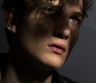 A close-up image of a man with blond hair and green eyes posing in a leather jacket. He’s staring into the distance as shadows cover parts of his face.