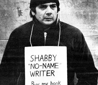 A black and white picture of Crad Kilodney — a middle-aged, gruff-looking white man in his 30s — in a toque and winter coat, wearing a sign around his neck that reads ‘Shabby ‘No-Name’ Writer — Buy my book — $2.’