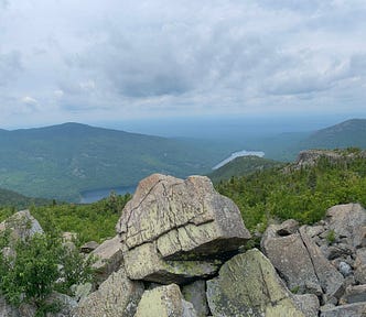 Mountaintop view with large boulders in the foreground and green mountain peaks in the background. In the middle, below, you can see a small lake peeking out. The sky is hazy with clouds and sun.