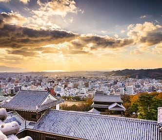 Sunset view over Matsuyama City from the rooftops of Matsuyama Castle, with golden light spilling across the skyline and the Seto Inland Sea glowing in the distance.
