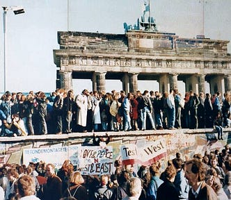 Color photograph of many people standing at the Bradenburg Gate at the Berlin Wall while a crowd below holds signs and banners.