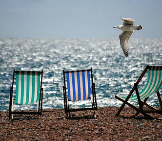 Three-green and blue stripy beach chairs on a pebbled beach with glittering waves in the background and a seagull flying by.